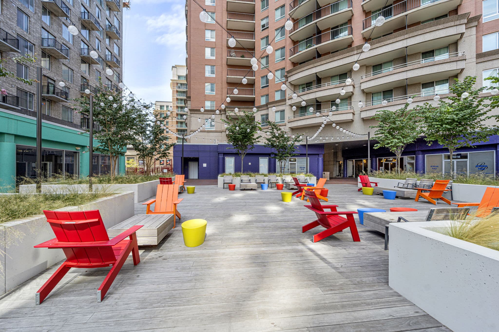 Outdoor urban courtyard with colorful chairs, benches, string lights, and planters, surrounded by tall apartment buildings and storefronts.